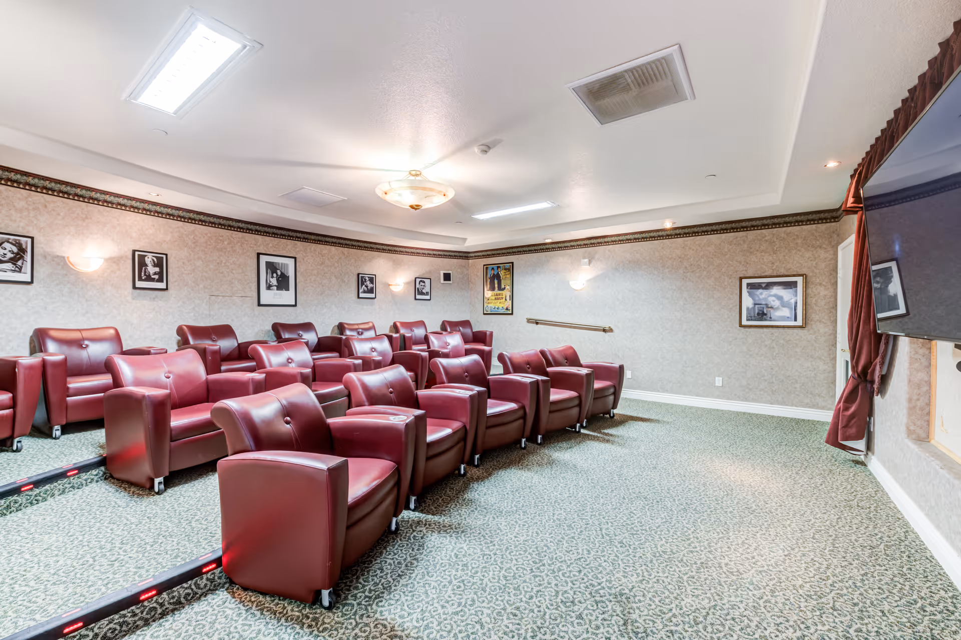 A small theater room with three rows of red leather recliner chairs arranged on carpeted steps facing a large flat-screen TV mounted on the wall. The walls are decorated with framed black and white photos and a movie poster. The room has soft lighting from ceiling fixtures and wall sconces.