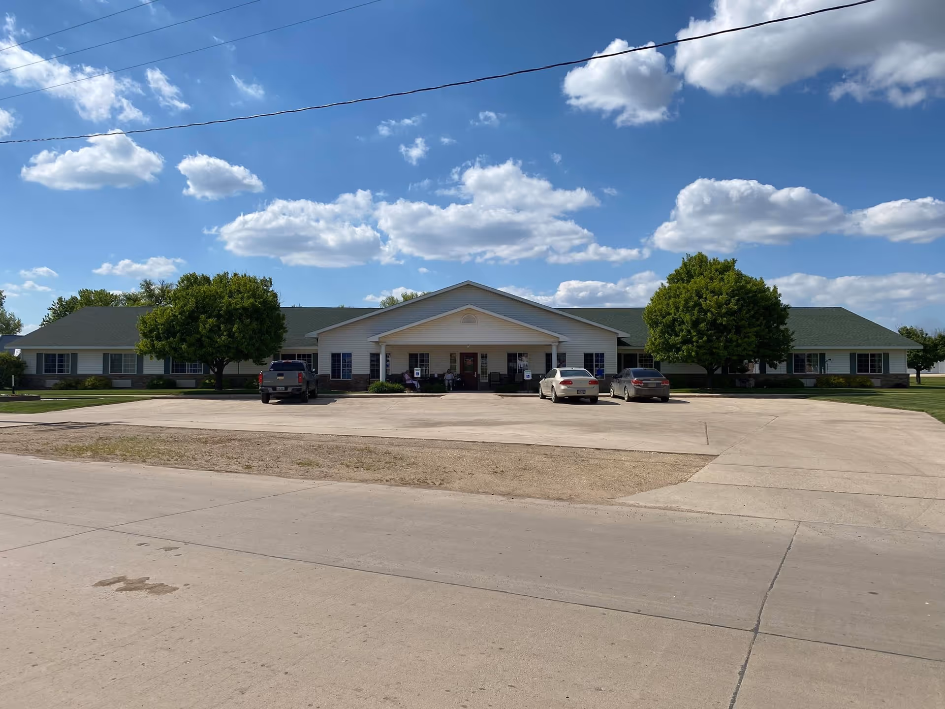 Front exterior view of Castlewood Assisted Living Center, a single-story building with a green roof, white siding, and a covered entrance. There are three vehicles parked in front and two large trees on either side of the entrance. The sky is blue with scattered clouds.