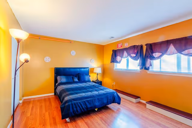 A bedroom with mustard yellow walls and wooden flooring. It features a black bed with dark blue bedding and pillows, a black nightstand with a lamp, a floor lamp with two lights, and two windows with dark purple curtains. There is an exit sign above the windows.