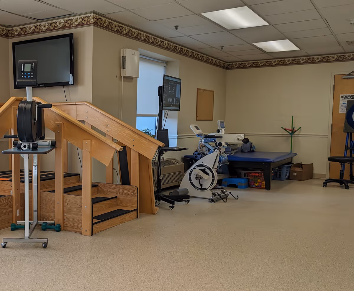 A rehabilitation or physical therapy room with wooden stairs for practice, an exercise bike, a therapy table, a wall-mounted TV, and various therapy equipment in a room with beige walls and a patterned border near the ceiling.