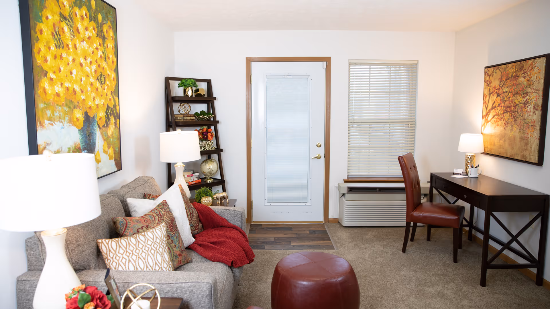A cozy living room with a gray sofa adorned with patterned pillows and a red throw blanket. There are two white lamps on either side of the sofa. A dark wooden ladder shelf with decorative items is placed near a white door with a window. To the right, there is a dark wooden desk with a red leather chair and a table lamp. The room has beige carpet flooring and two large paintings on the walls, one with yellow flowers and the other with an autumn tree scene.
