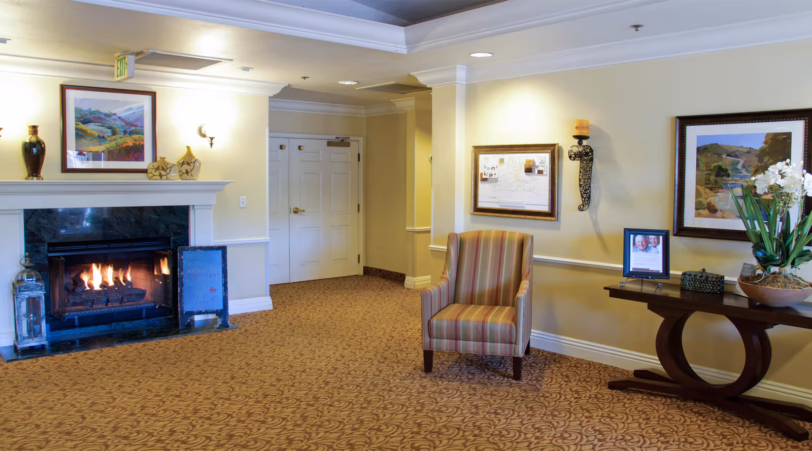 Lobby-style seating area with a lit fireplace, a striped armchair, a console table with a plant and framed artwork on the walls.