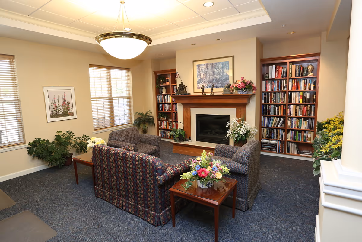 A cozy living room area in a retirement community featuring a fireplace with a wooden mantel, two bookshelves filled with books on either side, and a framed picture above the fireplace. The room has three upholstered chairs and a sofa arranged around a wooden coffee table with a floral arrangement. There are windows with blinds, potted plants, and a ceiling light fixture.