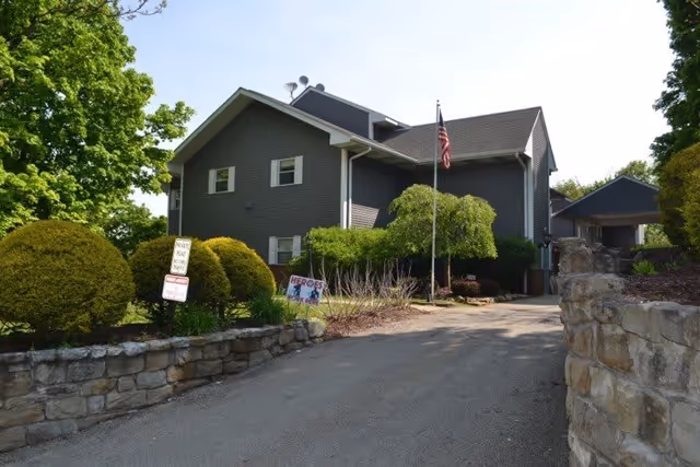Exterior view of a two-story gray building with white trim, surrounded by green trees and bushes. A stone wall lines the driveway leading up to the building. An American flag is mounted on a pole near the entrance, and a sign with the word 'HEROES' is visible in the garden area.