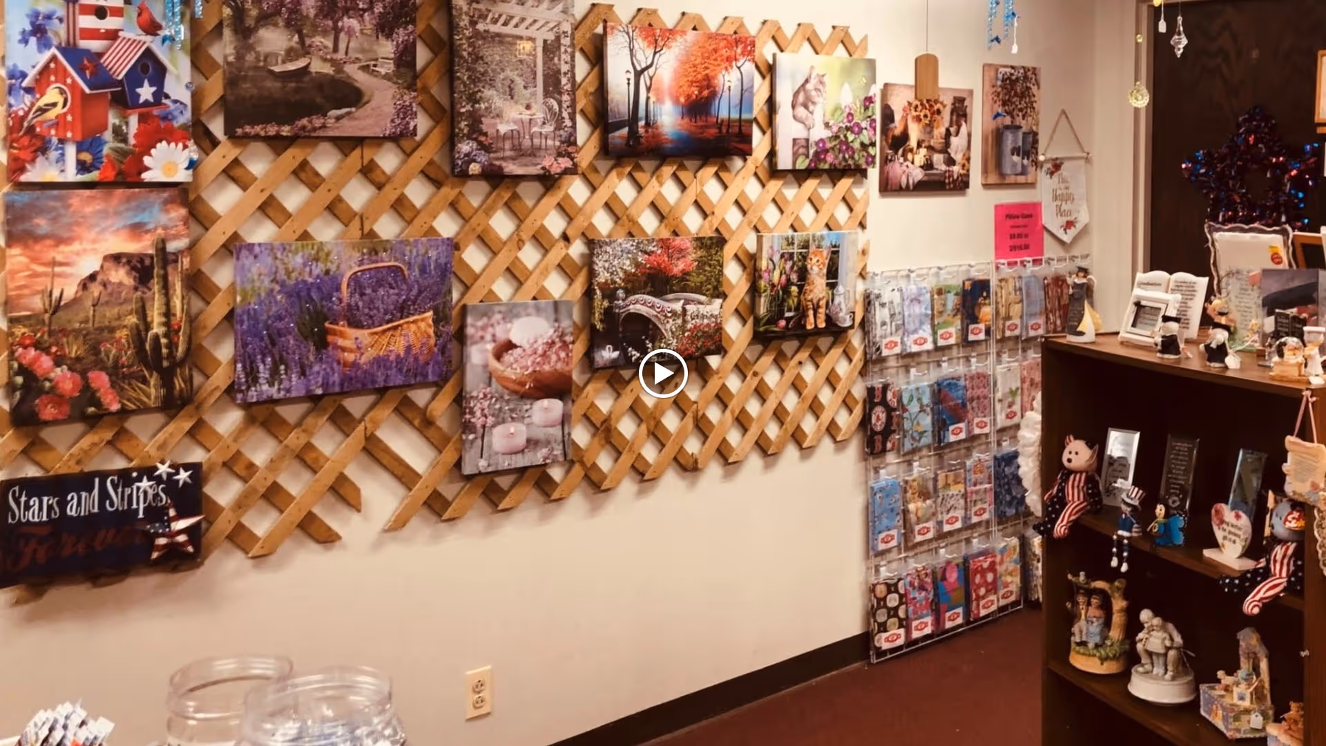 Interior view of a room with a lattice wall decorated with various colorful paintings and pictures. To the right, there is a display rack with greeting cards and a wooden shelf filled with small decorative figurines and items. The room has a cozy and welcoming atmosphere.