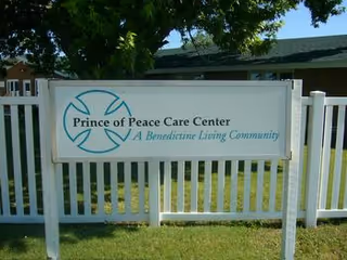 White fence with a sign that reads 'Prince of Peace Care Center A Benedictine Living Community' in front of a building and green lawn with a tree providing shade.