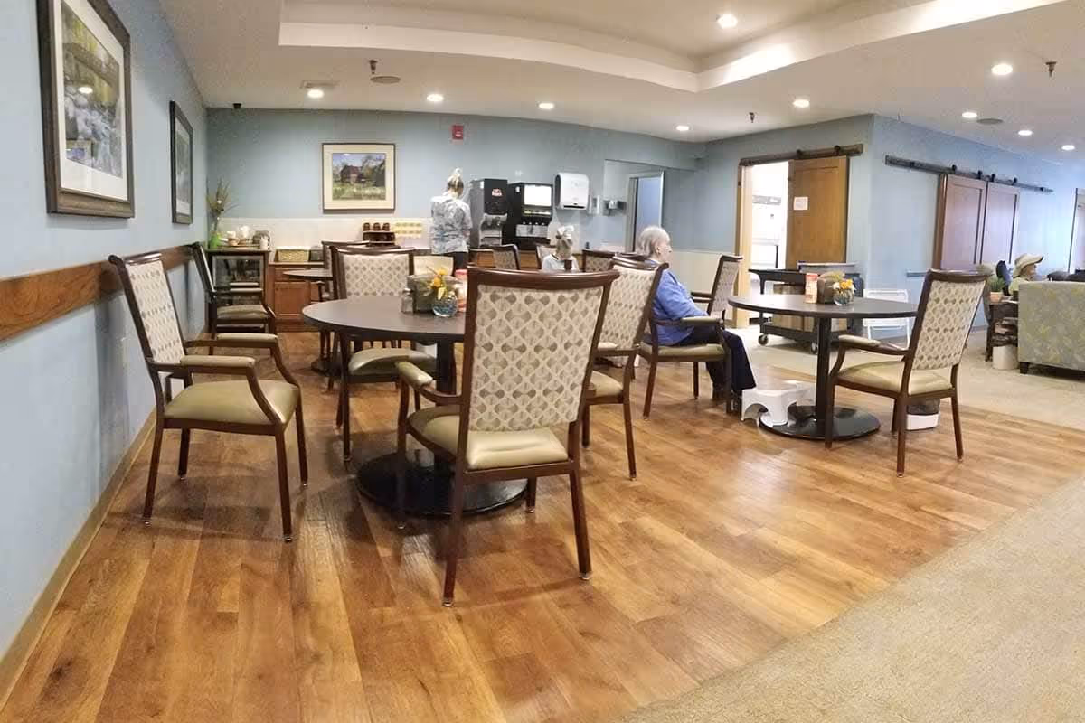 A dining area in a senior living facility with several round tables and cushioned chairs. A few elderly residents are seated at the tables, and a staff member is seen near a beverage station against the wall. The room has wooden flooring and light blue walls with framed artwork.