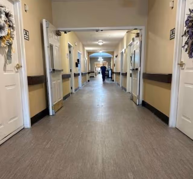 A long hallway in an assisted living facility with beige walls, multiple white doors on both sides, some decorated with wreaths, and a person walking in the distance. The floor is covered with light brown tiles and the hallway is well-lit with ceiling lights.