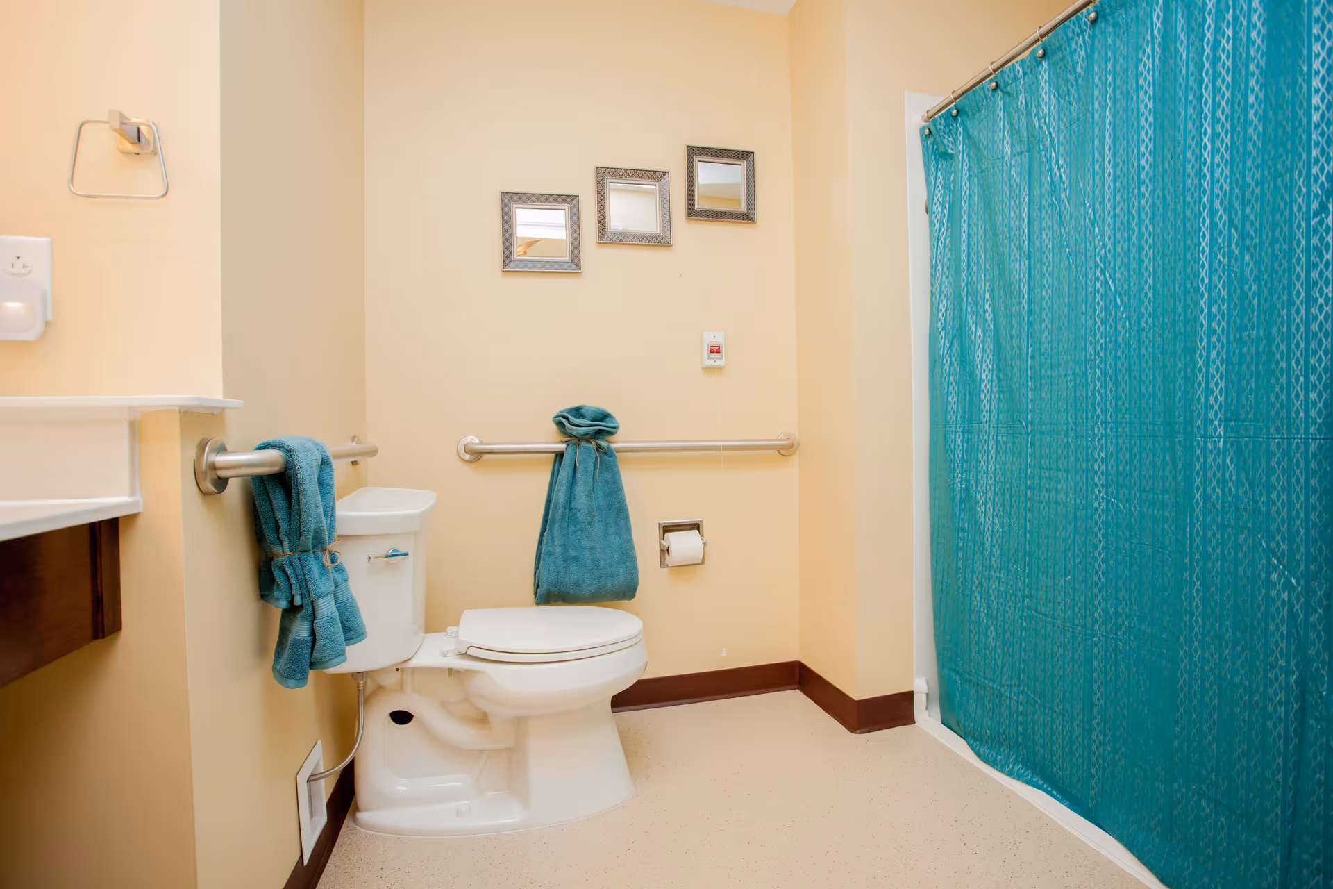 A clean bathroom with beige walls and floor, featuring a white toilet with teal towels hanging on grab bars beside and behind it. There is a teal shower curtain on the right side and three small decorative mirrors on the wall above the toilet.