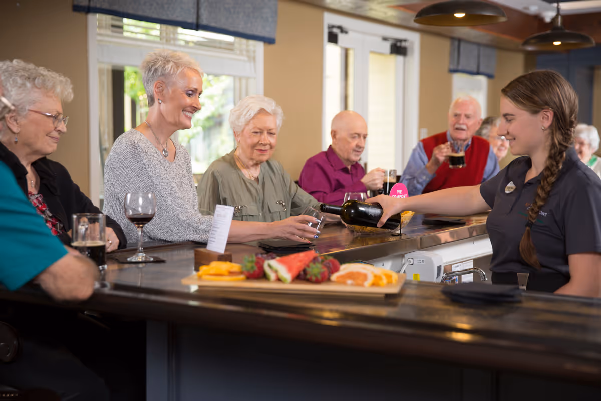 A group of elderly people sitting at a bar counter inside a facility, being served drinks by a smiling female staff member. There is a platter of sliced fruits on the counter and large windows in the background letting in natural light.