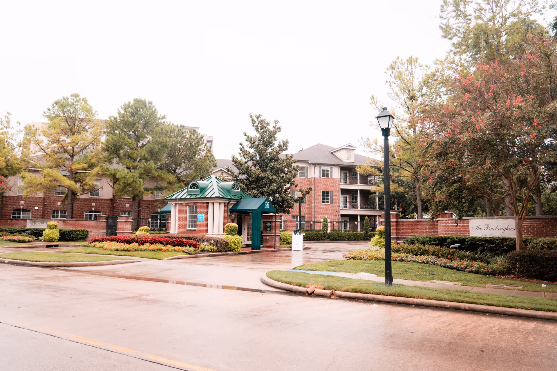 Entrance to The Buckingham senior living facility featuring a brick guardhouse with a green roof, surrounded by landscaped flower beds and trees, with the main building visible in the background.