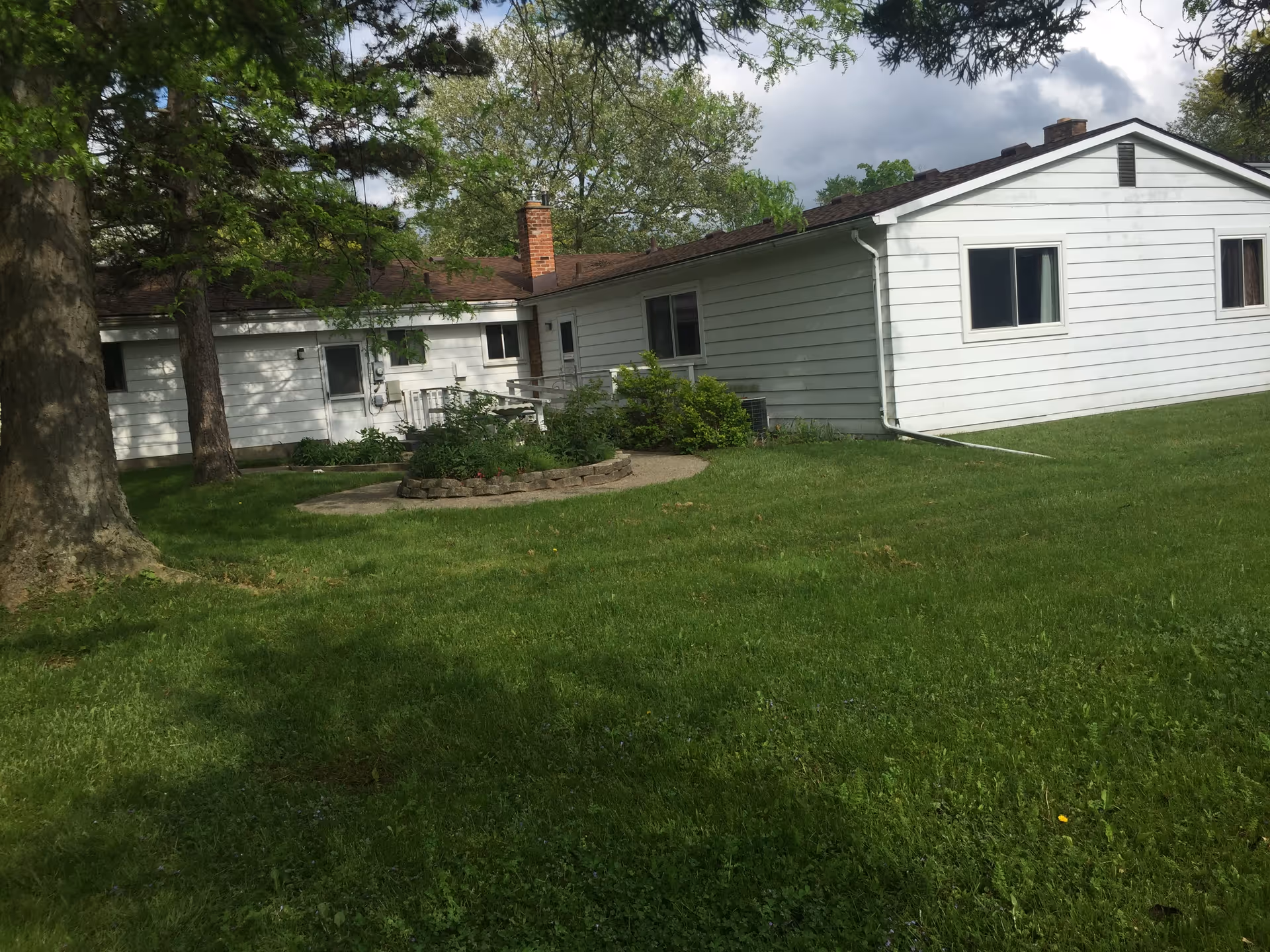 Exterior view of a single-story white building with a brown roof, surrounded by green grass and trees under a partly cloudy sky. There is a small circular garden bed with plants near the building.