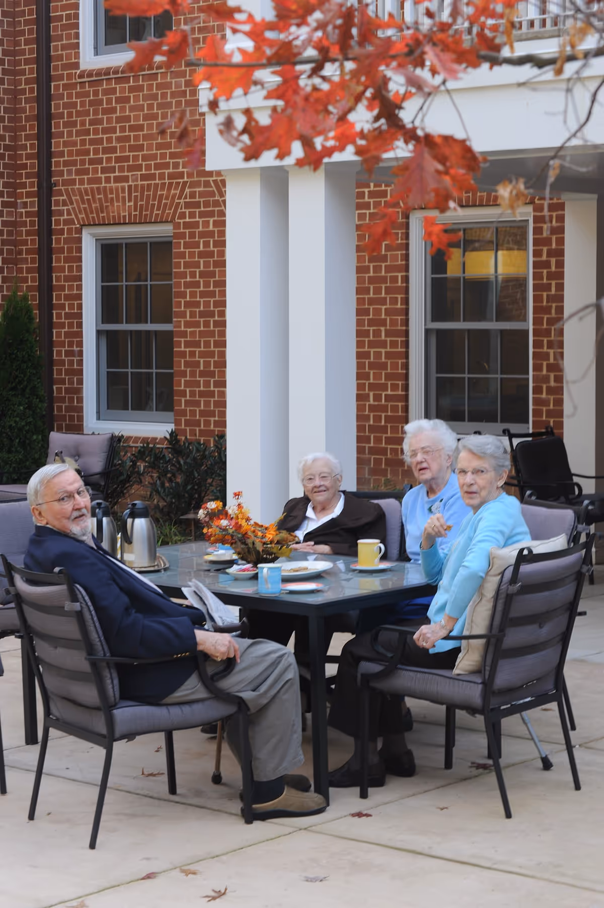 Four elderly people sitting around a glass outdoor table with chairs on a patio. They are enjoying a casual gathering with coffee cups and a floral centerpiece. The background shows a brick building with white pillars and windows, and some autumn leaves hanging above.