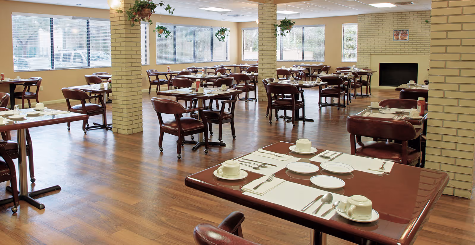 A spacious dining room with multiple tables set for meals, each table having white plates, cups, and silverware. The room has wooden flooring, beige brick pillars, large windows with blinds, and hanging plants near the ceiling. The chairs are cushioned and upholstered in dark red leather.