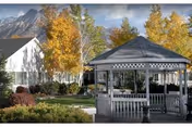Outdoor view of a senior living facility area featuring a white gazebo with lattice trim surrounded by green grass, shrubs, and trees with autumn foliage. In the background, there is a white building and mountains under a partly cloudy sky.