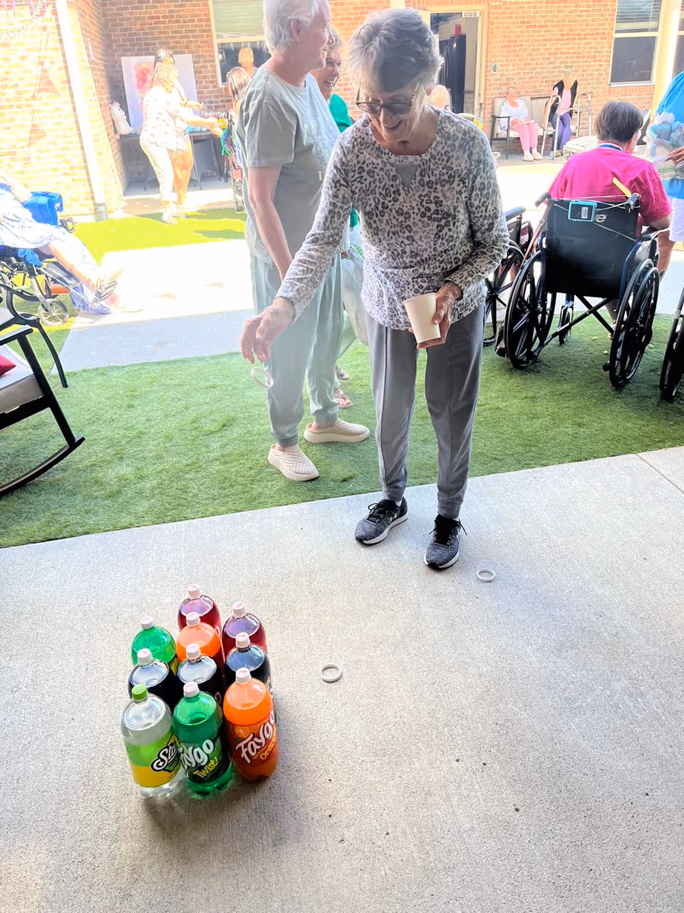 An elderly woman wearing glasses and a leopard print top is playing a ring toss game outdoors on a concrete surface. Several large soda bottles are arranged like bowling pins on the ground. Other elderly people, some in wheelchairs, are visible in the background near a brick building and artificial grass area.