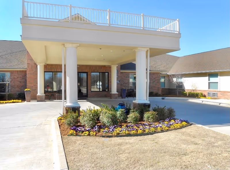Entrance of a senior living facility with a covered drop-off area supported by white columns, surrounded by a flower bed with yellow and purple flowers, and a clear blue sky above.