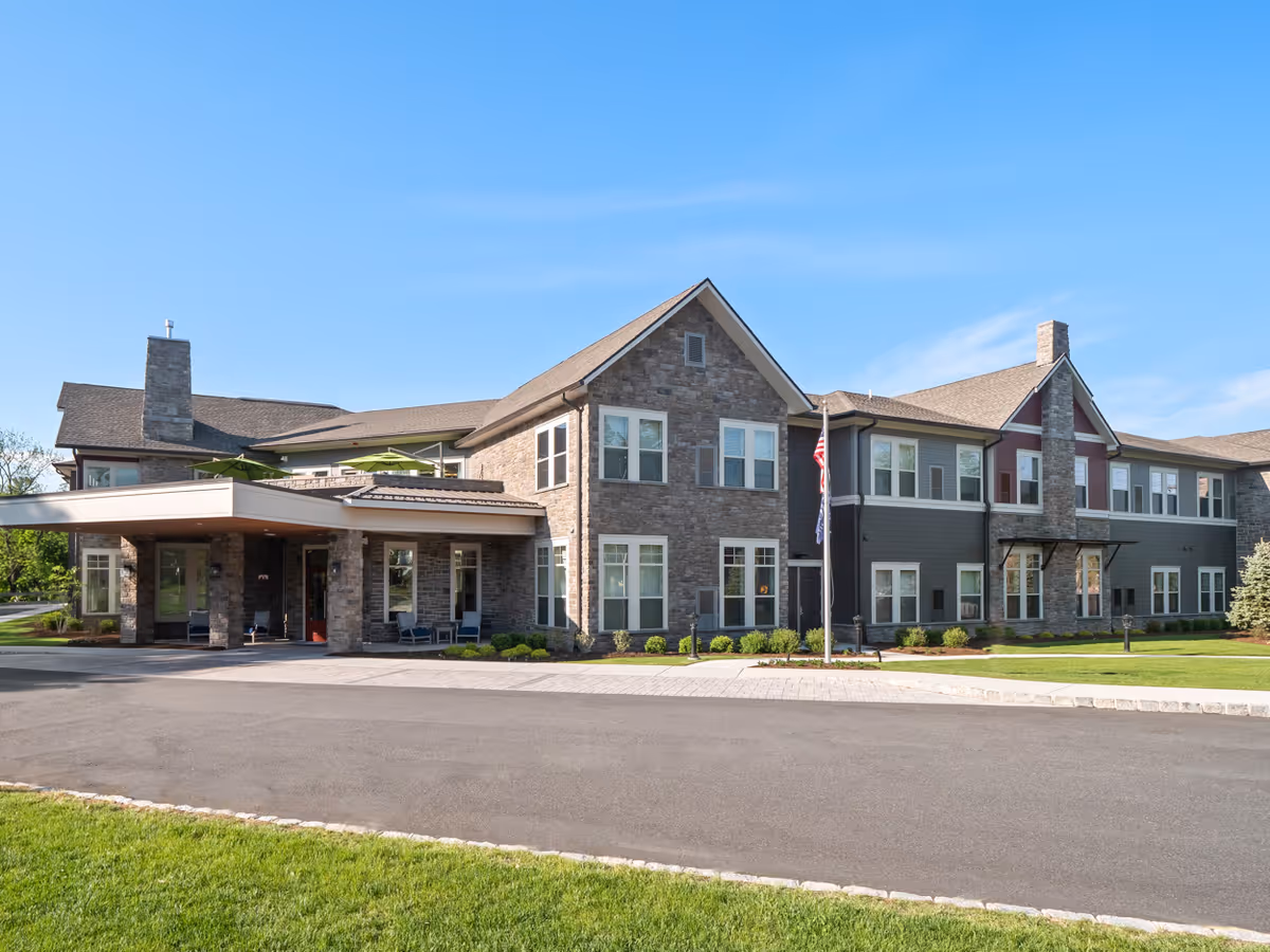 Exterior view of The Bristal Assisted Living at Waldwick building with stone and siding facade, multiple windows, two chimneys, and a covered entrance. The building is surrounded by a paved driveway, green lawn, and clear blue sky.