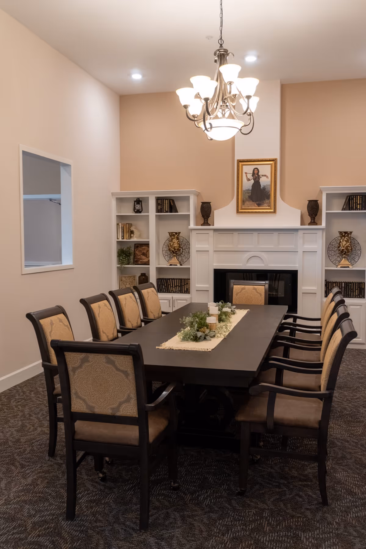 A formal dining room with a long dark wooden table surrounded by eight upholstered chairs. The table is decorated with a beige runner and greenery with candles. Behind the table is a white fireplace with built-in shelves on either side holding decorative items and books. Above the fireplace hangs a framed painting of a woman. A chandelier with multiple lights hangs from the ceiling, illuminating the room.