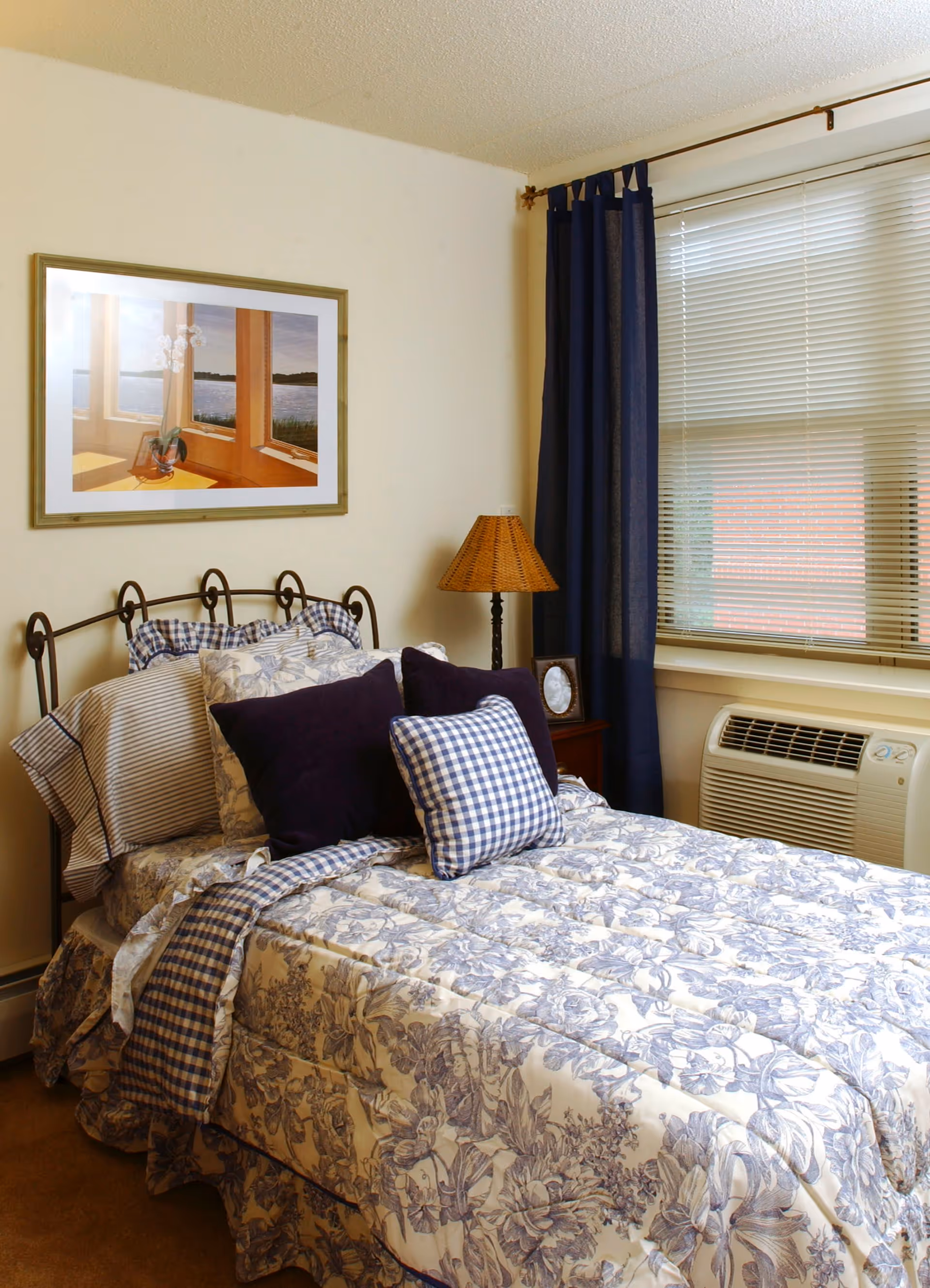 A cozy bedroom with a single bed featuring floral and checkered bedding in shades of blue and white. The bed has multiple pillows, including dark blue and checkered ones. Next to the bed is a wooden nightstand with a wicker lampshade and a small framed photo. A window with closed blinds and dark blue curtains is on the right side of the room. A framed picture of a window with a view of water and flowers hangs on the wall above the bed.