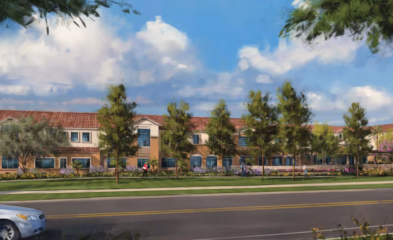 Front exterior of a two-story senior living building with a red tile roof, trees, and landscaped lawns along a street.