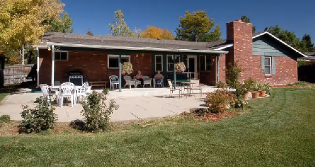 A single-story brick house with a covered patio area. The patio has several white plastic chairs and tables, with a few people sitting and relaxing. The house is surrounded by a well-maintained lawn and some small bushes. Trees with autumn-colored leaves are visible in the background under a clear blue sky.