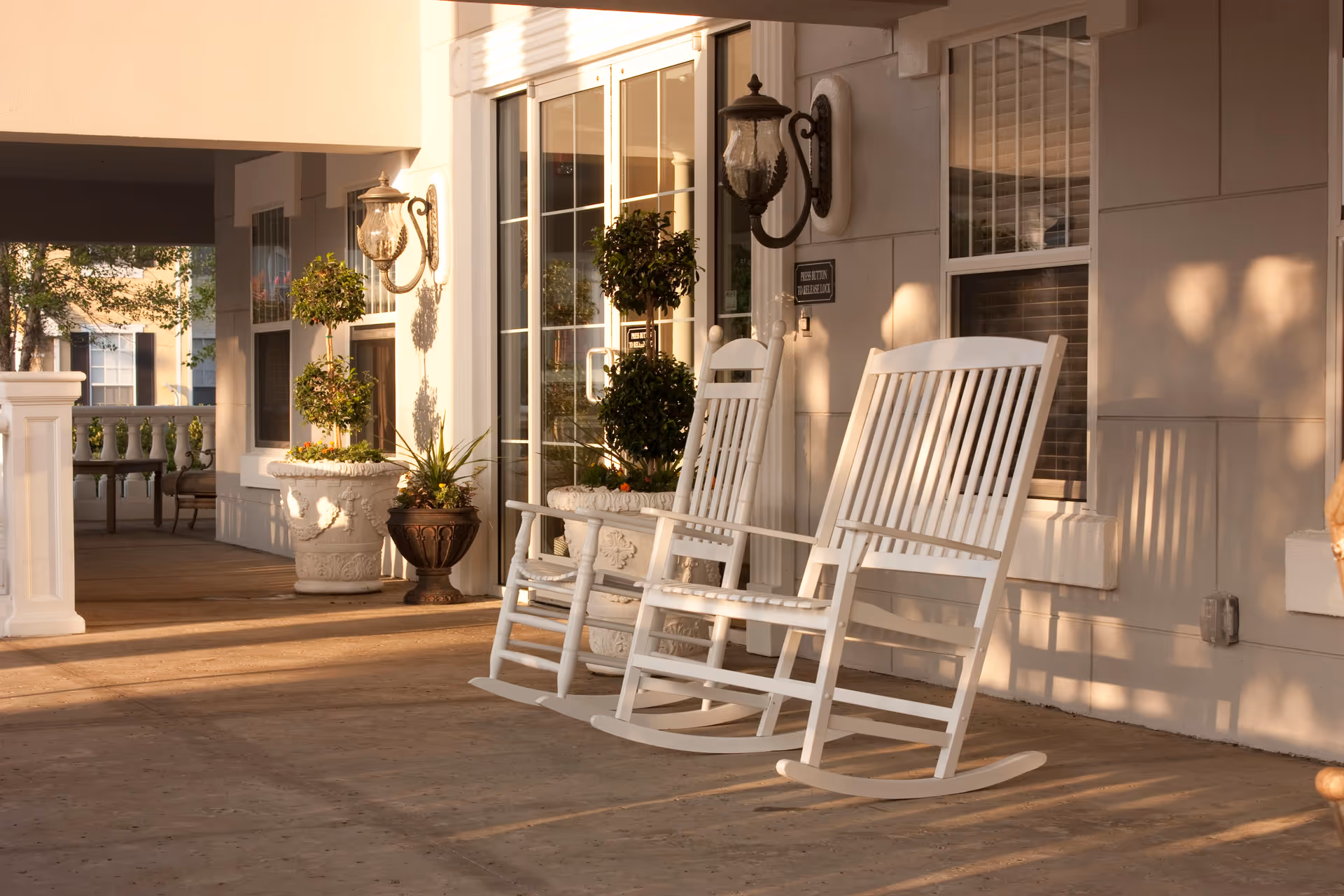 Two white wooden rocking chairs on a covered porch outside a building with large windows and decorative potted plants, bathed in warm sunlight.