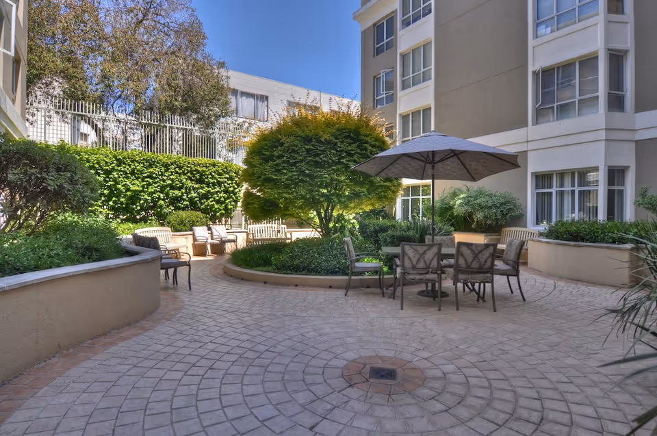 Outdoor courtyard area at Ivy Park at Cathedral Hill featuring a circular paved patio with a table, chairs, and a large umbrella. The space is surrounded by greenery including bushes and a tree, with the building's windows visible in the background under a clear blue sky.