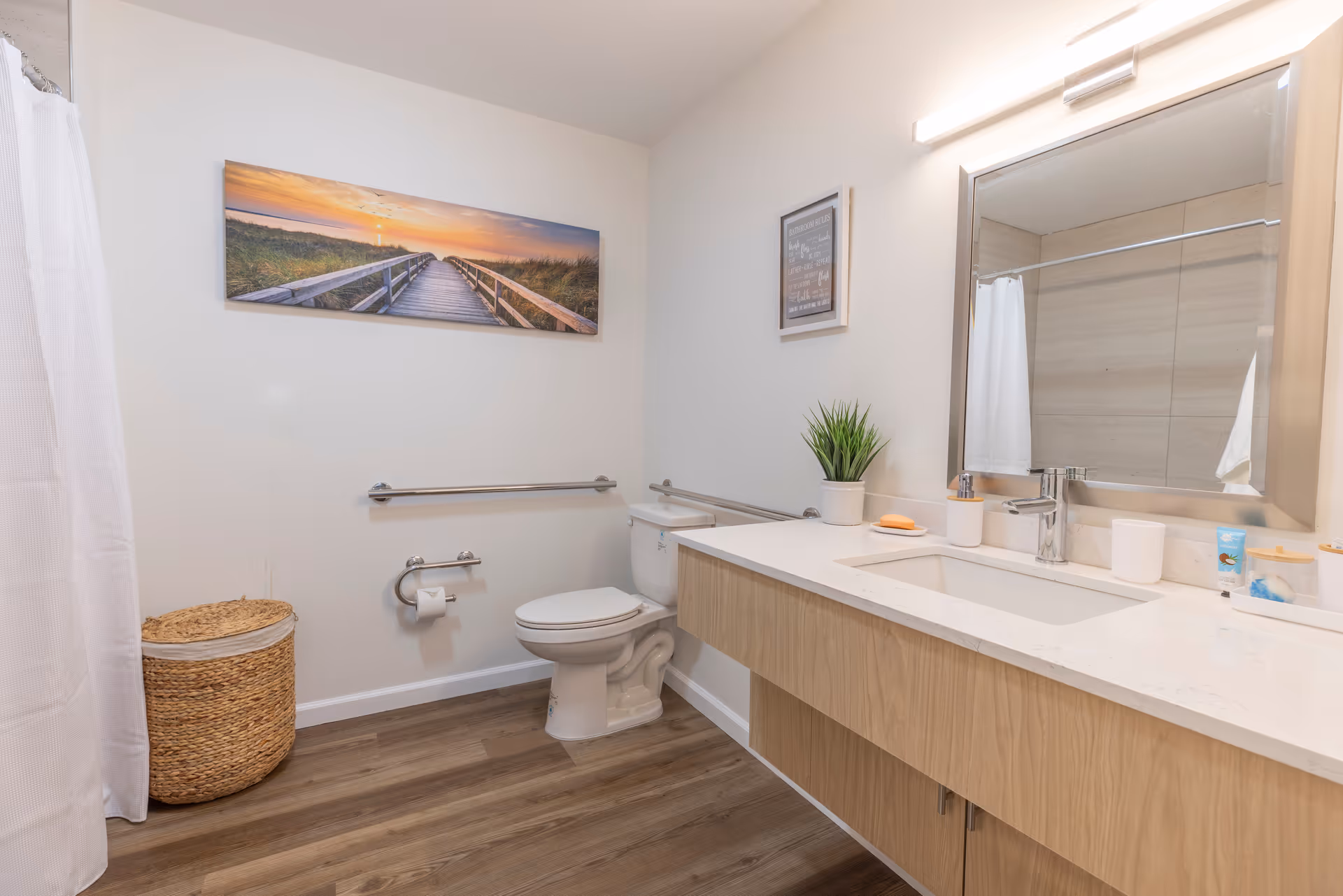 Bright accessible bathroom with a long vanity and sink, toilet with grab bars, shower curtain, and wall art.