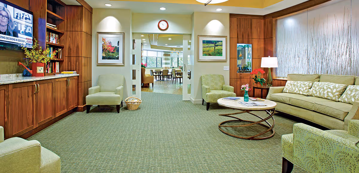 A cozy living room area in Asbury Solomons Island featuring a green sofa with patterned cushions, several green armchairs, a round coffee table with a vase of flowers and magazines, wooden built-in shelves with books and decorative items, a wall-mounted TV showing a news program, framed artwork on the walls, and a view into a dining area through an open doorway.