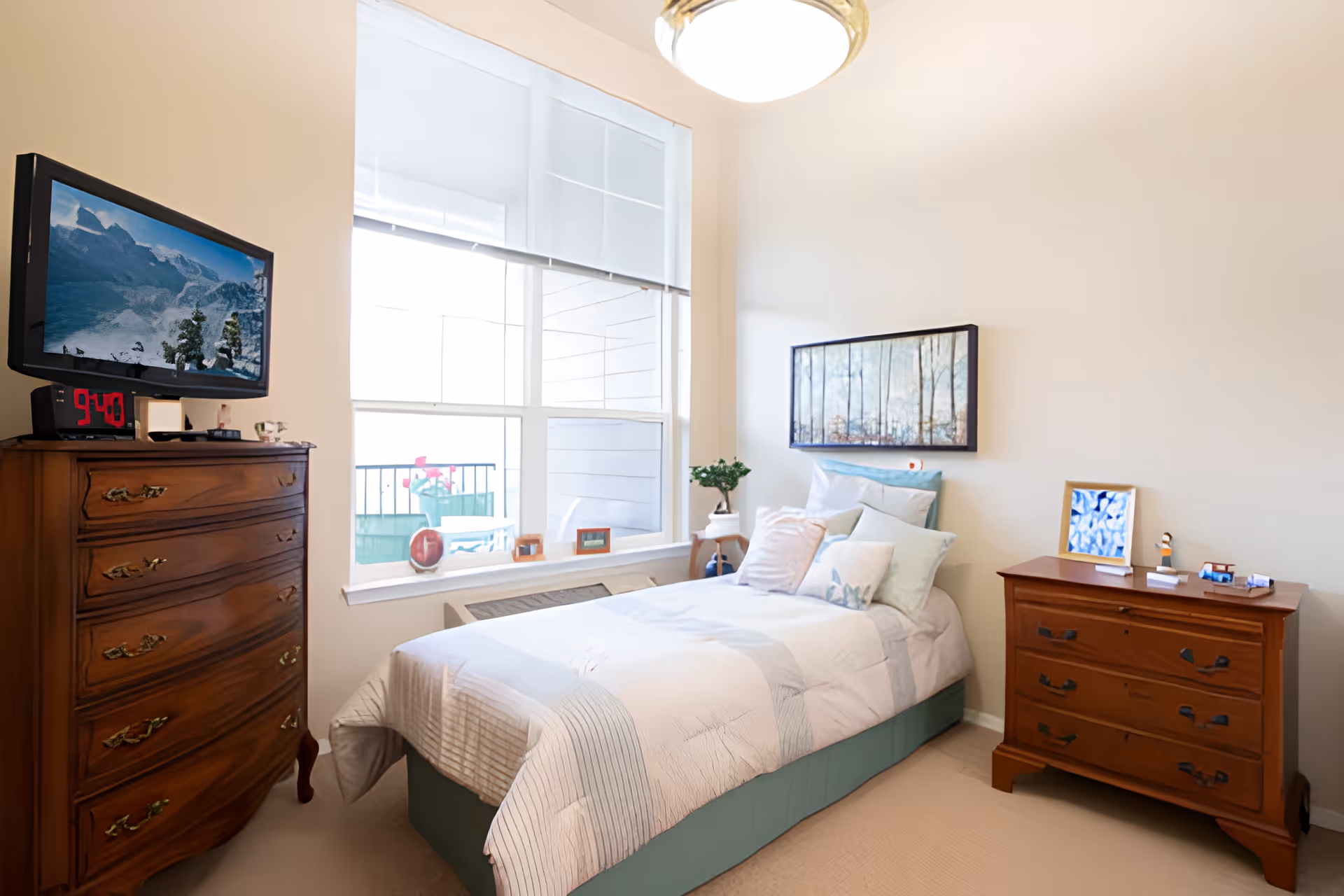 A bright and cozy bedroom in a senior living facility featuring a single bed with multiple pillows and a light-colored comforter. There is a wooden dresser with a digital clock and a TV mounted on the wall above it on the left side. On the right side, there is another wooden dresser with decorative items and a framed picture. A large window with white blinds lets in natural light, and a framed artwork of trees hangs on the wall above the bed.