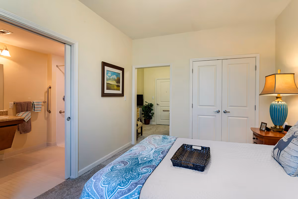 A bedroom in an assisted living facility with a bed covered in a white quilt and a blue patterned throw. A wicker tray is placed on the bed. To the right, there is a wooden nightstand with a blue lamp and some small framed pictures. The room has beige walls and carpeted floor. A doorway on the left leads to a bathroom with a sink, mirror, and towel racks with towels. Another doorway in the center leads to a hallway with a plant and a door visible.