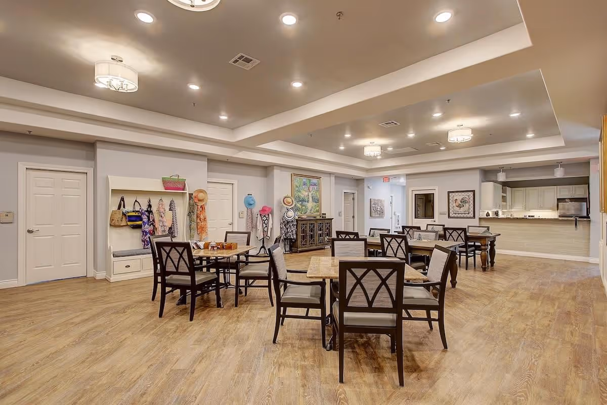 Open communal dining room in a senior living facility with several tables and chairs, a coat rack, and a serving counter in the back.