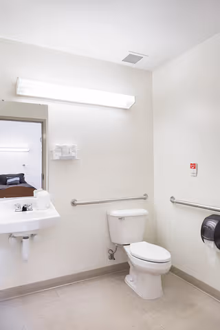 A clean, accessible bathroom with a white toilet, a white sink with a faucet, grab bars on the walls, a wall-mounted paper towel dispenser, and a large rectangular light fixture above a mirror. The bathroom has light-colored walls and flooring, and a doorway leading to a bedroom is visible in the reflection of the mirror.