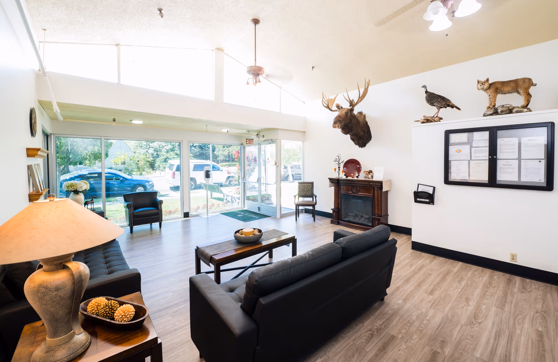 A bright and spacious living room area with large windows and glass doors letting in natural light. The room features black leather sofas, a wooden coffee table with decorative items, a side table with a lamp, and a fireplace with mounted animal heads above it, including a moose, a bird, and a bobcat. There is a bulletin board on the wall and a couple of chairs near the entrance.