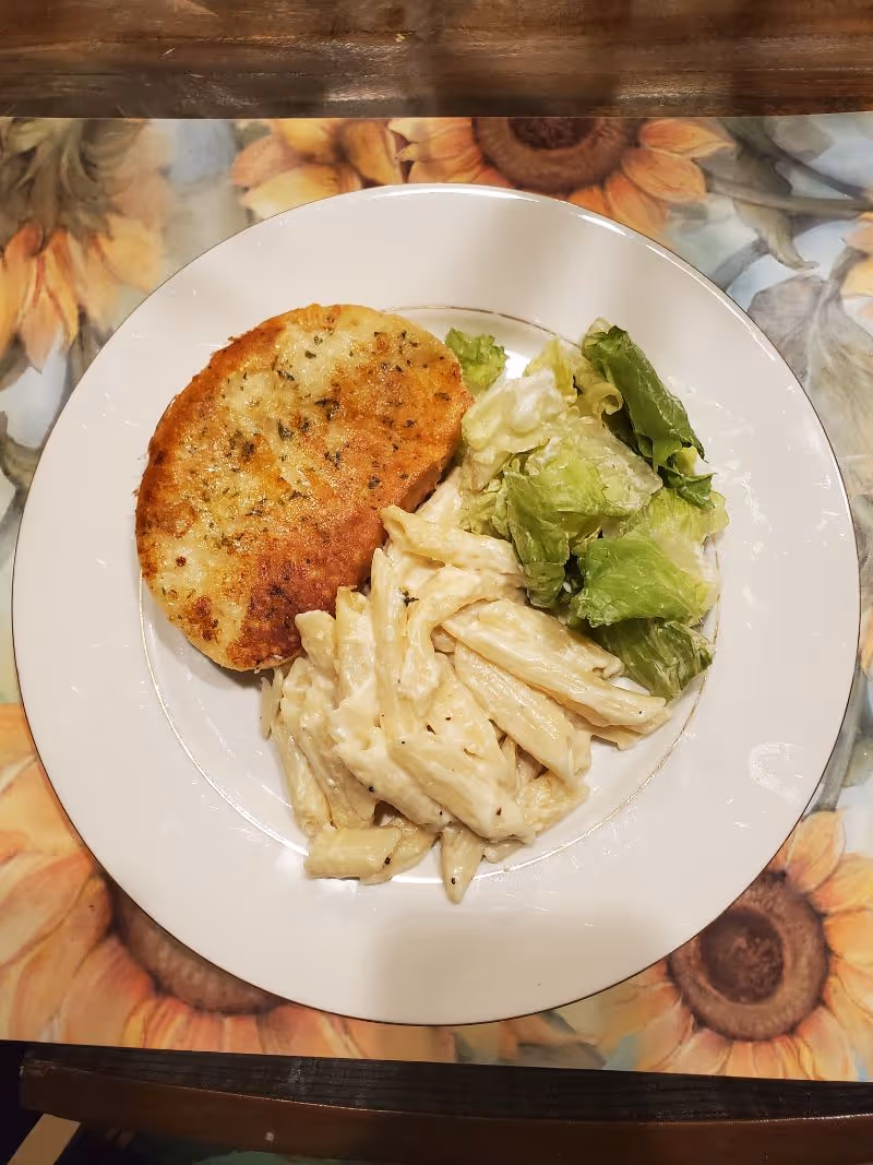 A white plate with a serving of creamy penne pasta, a portion of green leafy salad, and a piece of breaded and baked chicken, placed on a sunflower-themed placemat on a wooden table.
