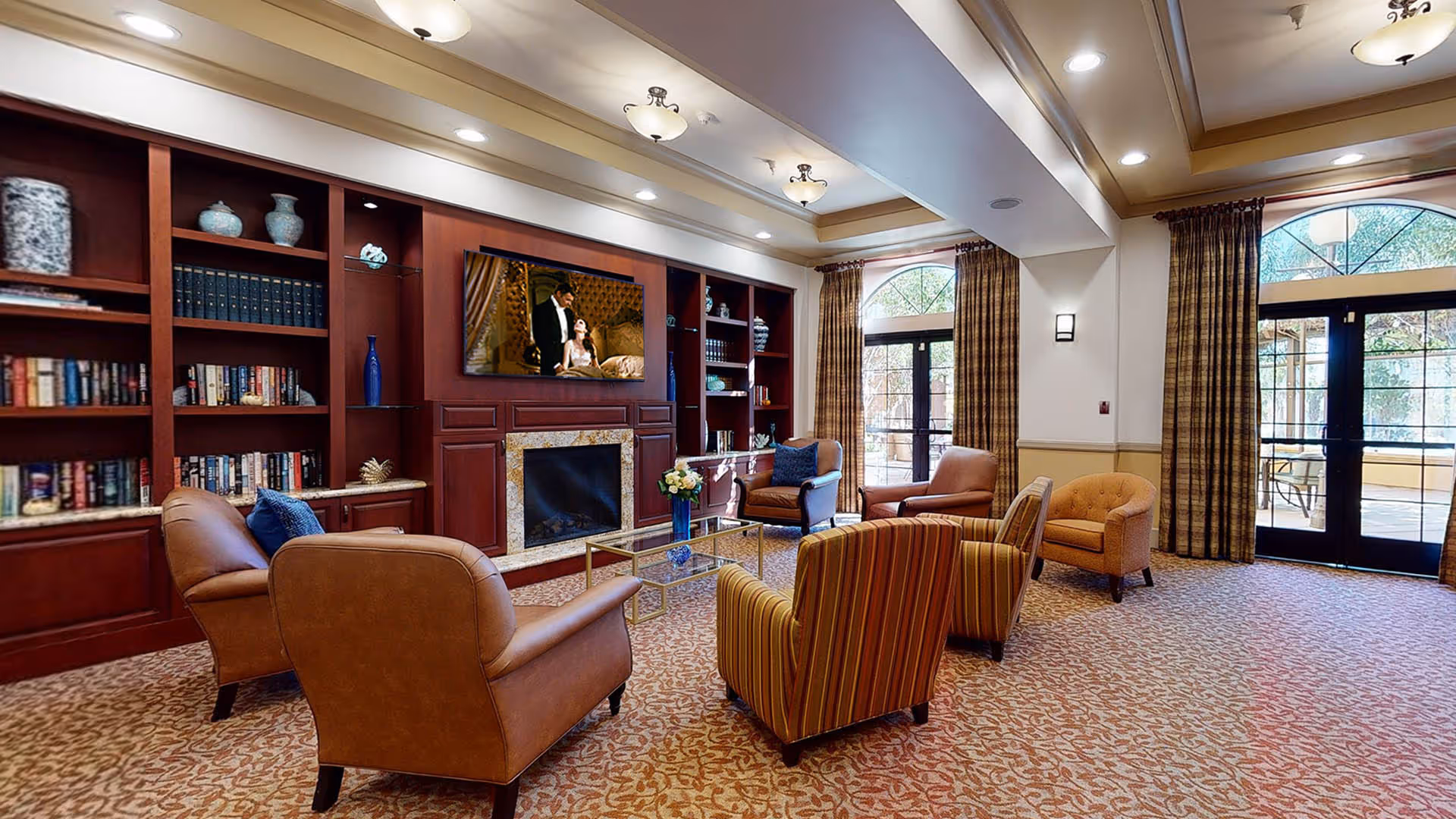 A cozy living room with a patterned carpet, several upholstered armchairs arranged around a glass coffee table with a blue vase of flowers, a built-in wooden bookshelf with decorative items and books, a fireplace with a mounted flat-screen TV above it, and large windows with arched tops and plaid curtains letting in natural light.
