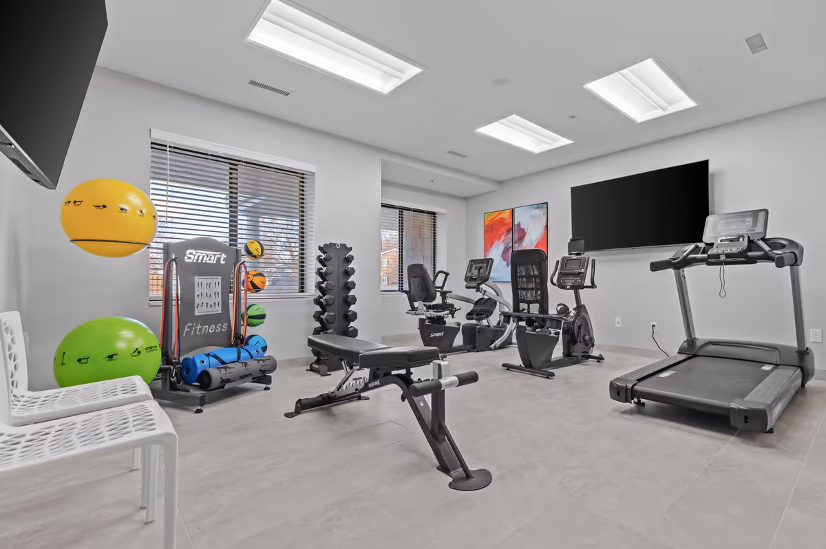 A bright fitness room with exercise equipment including a treadmill, two recumbent bikes, a weight bench, dumbbells, exercise balls, and yoga mats. The room has large windows with blinds, white walls, tiled floor, and two colorful abstract paintings on the wall. There is a large flat-screen TV mounted on the wall.