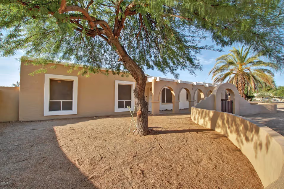 Exterior view of a single-story building with beige stucco walls and white window frames, featuring an arched walkway and a gated entrance. In the foreground, there is a tree with green foliage and a sandy ground area. A palm tree is visible in the background under a clear blue sky.