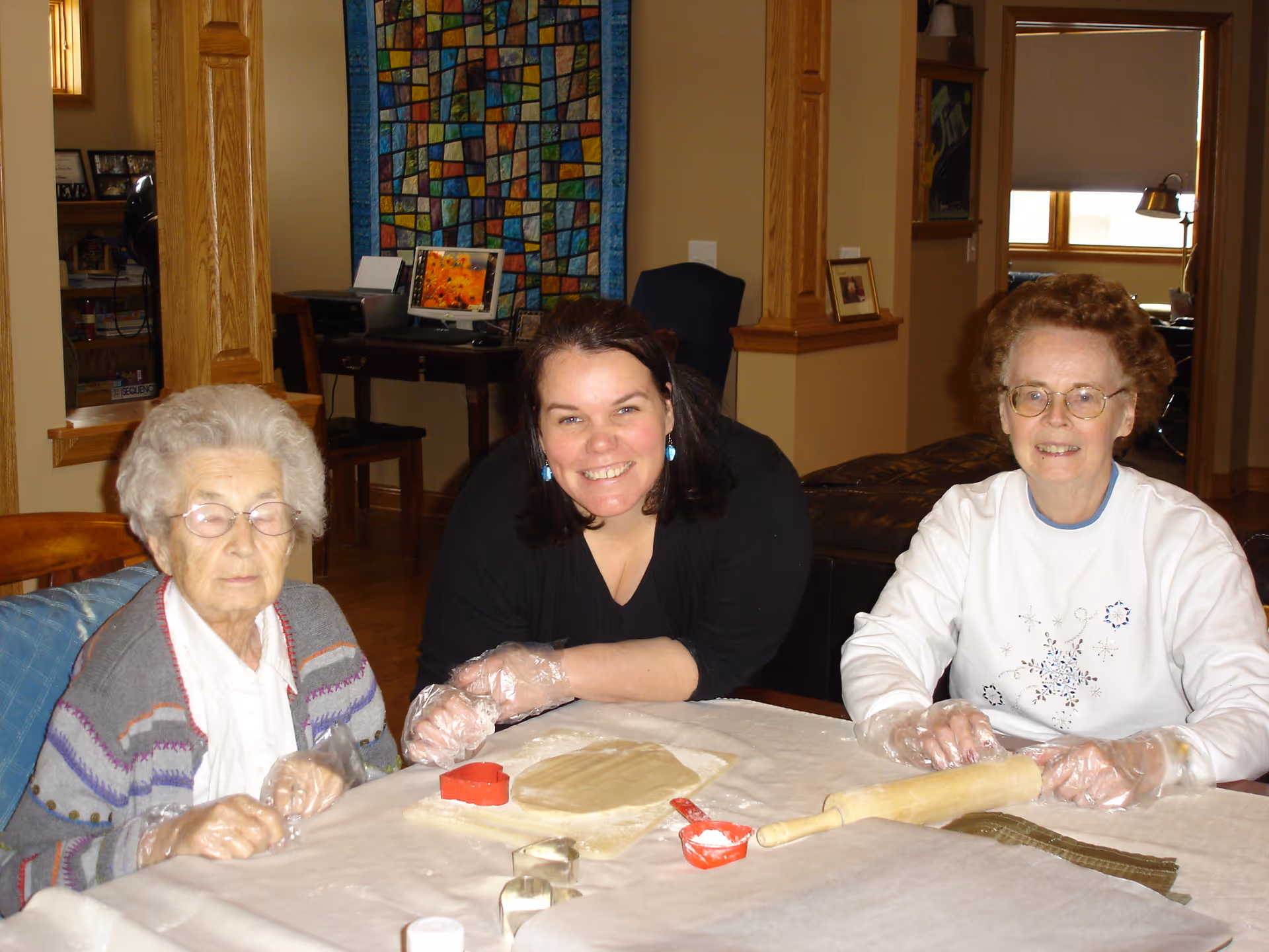 Three women sitting at a table covered with a white cloth, wearing plastic gloves and engaged in a baking activity with dough, cookie cutters, and a rolling pin. They are smiling and appear to be in a cozy indoor setting with wooden furniture and a colorful quilt hanging on the wall in the background.