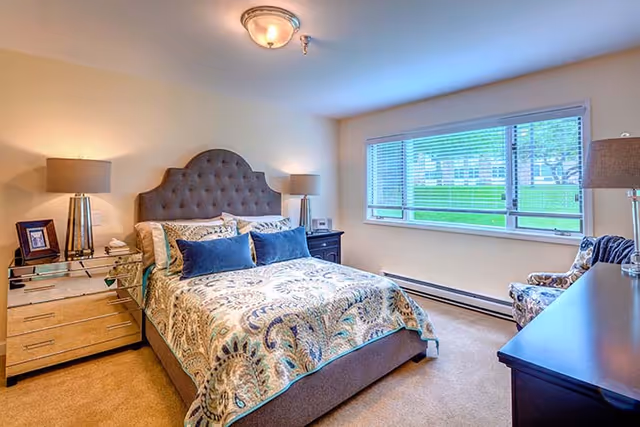 A well-lit bedroom featuring a large bed with a tufted headboard, patterned bedding, and multiple pillows. There are two bedside tables with lamps on each side of the bed. A large window with blinds lets in natural light, and a patterned armchair is placed near the window. A dark dresser with a lamp is also visible in the room.