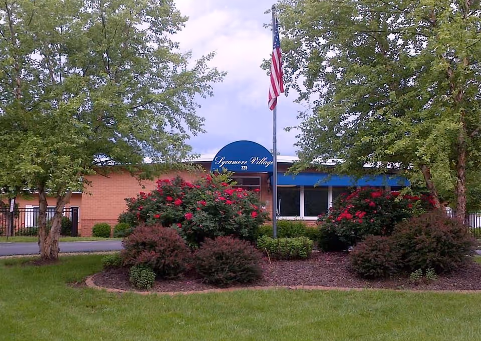 Exterior view of Sycamore Village Assisted Living building with a blue awning displaying the facility name and number 225. The foreground features a landscaped garden with green grass, bushes, and flowering plants, and an American flag on a flagpole is visible near the center.