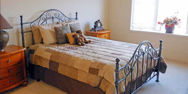 Sunlit bedroom with a metal-framed bed, quilted bedding, nightstands and a teddy bear on the bed.