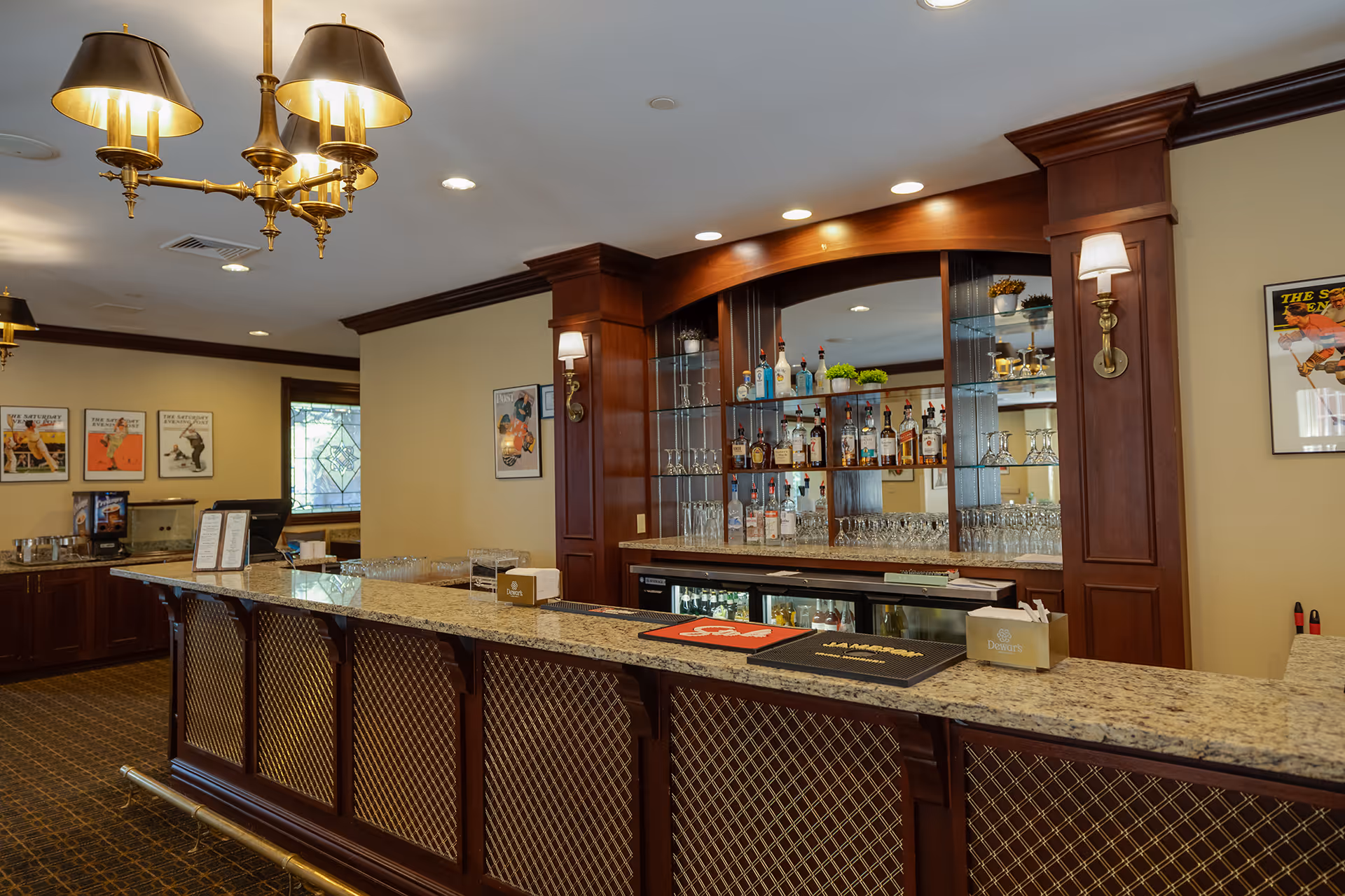 Interior view of a bar area in a senior living facility named Evergreen Woods, featuring a long granite countertop with a brass foot rail, wooden cabinetry with glass shelves displaying various bottles of liquor and glassware, warm lighting fixtures including wall sconces and a chandelier, and framed vintage posters on the walls.