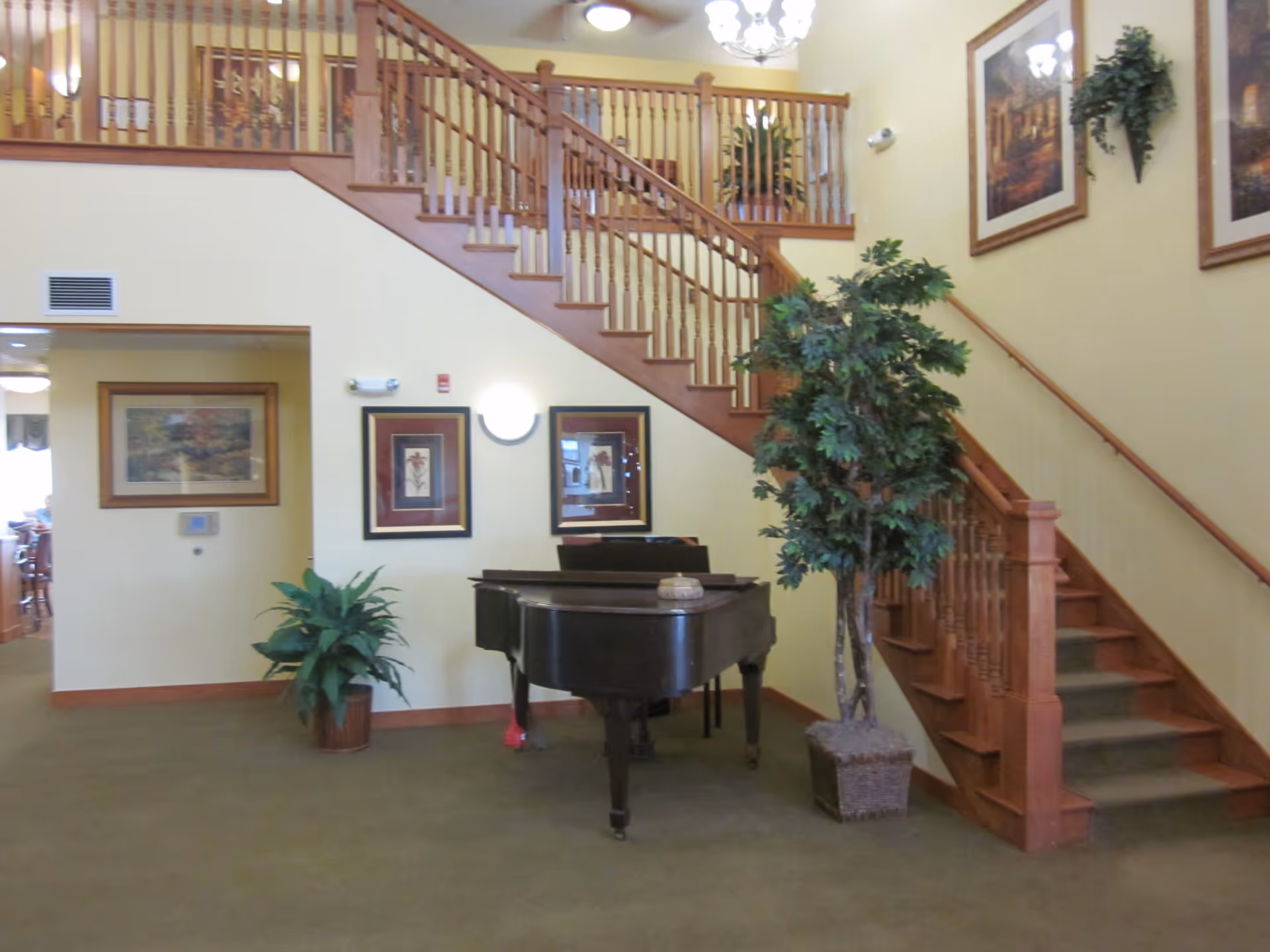 Interior view of a senior living facility showing a wooden staircase with a railing, a black grand piano, two potted plants, and framed artwork on the walls. The walls are painted light yellow and the floor is carpeted in green.
