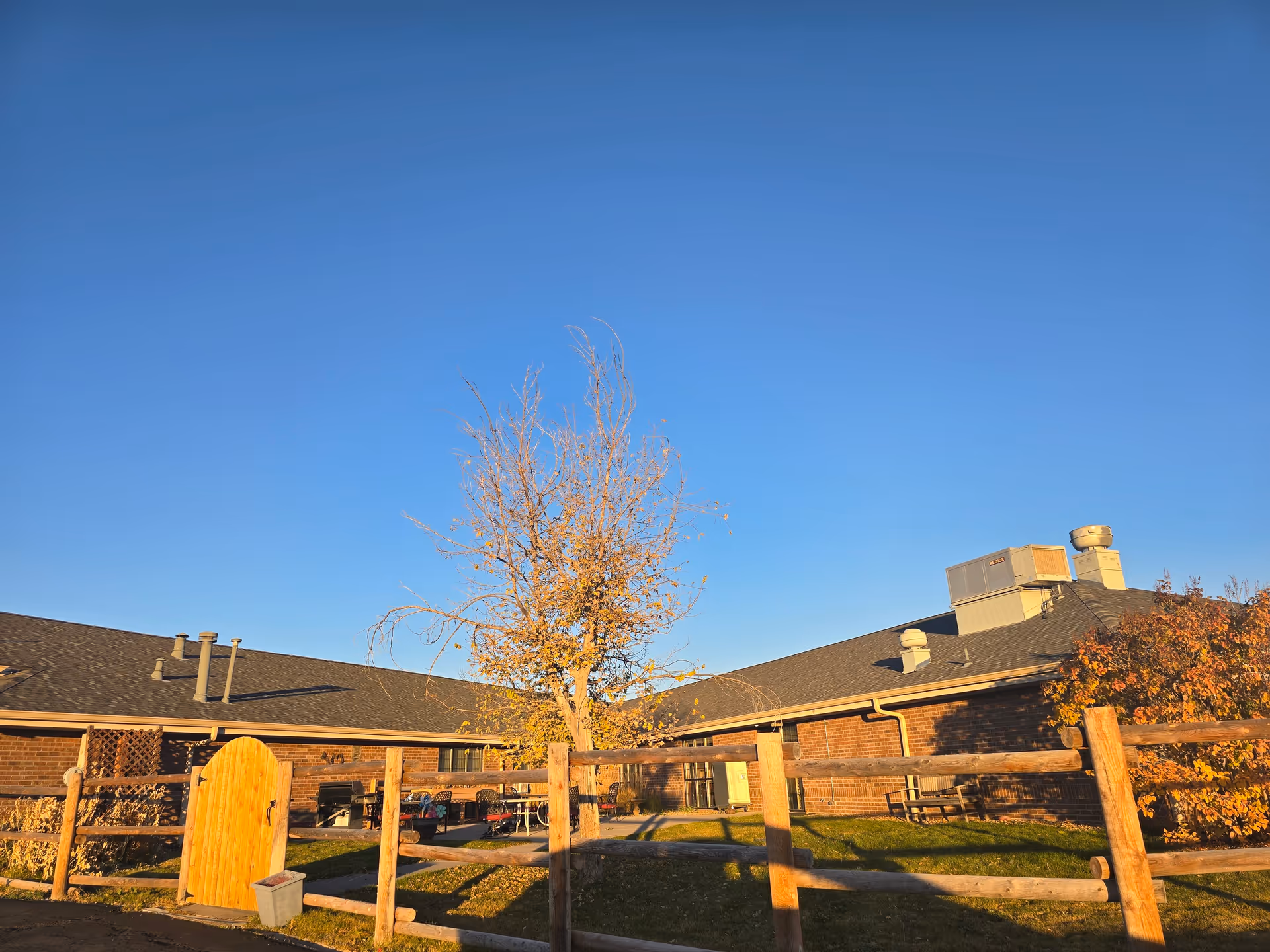 Exterior view of a single-story brick building with a wooden fence, a small yard and a leafless tree under a clear blue sky.