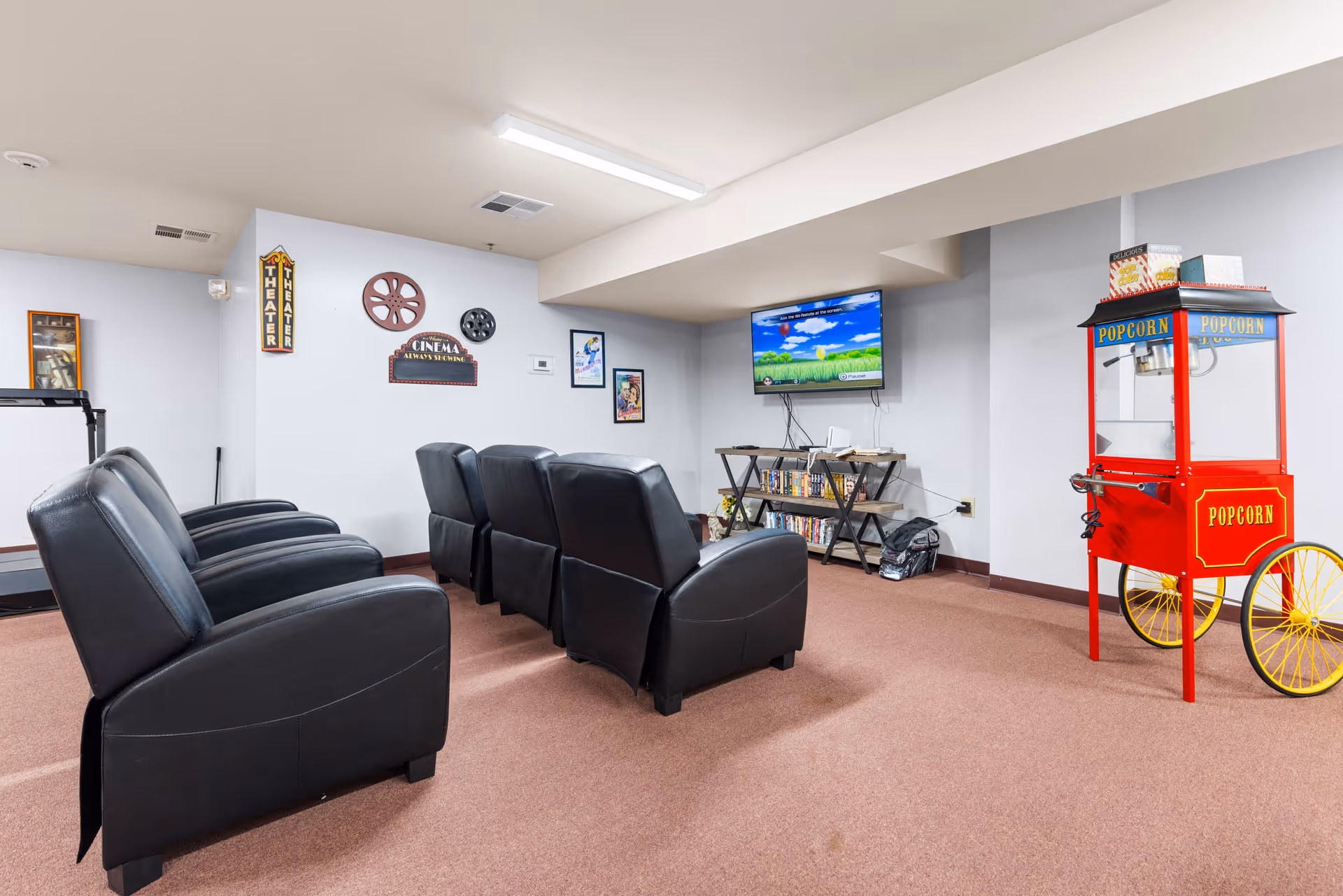 A cozy movie theater room in an assisted living facility with two rows of black leather recliner chairs facing a wall-mounted flat screen TV. The walls are decorated with cinema-themed signs and film reels. A red popcorn machine with yellow wheels is positioned on the right side of the room. The floor is carpeted in a light brown color.