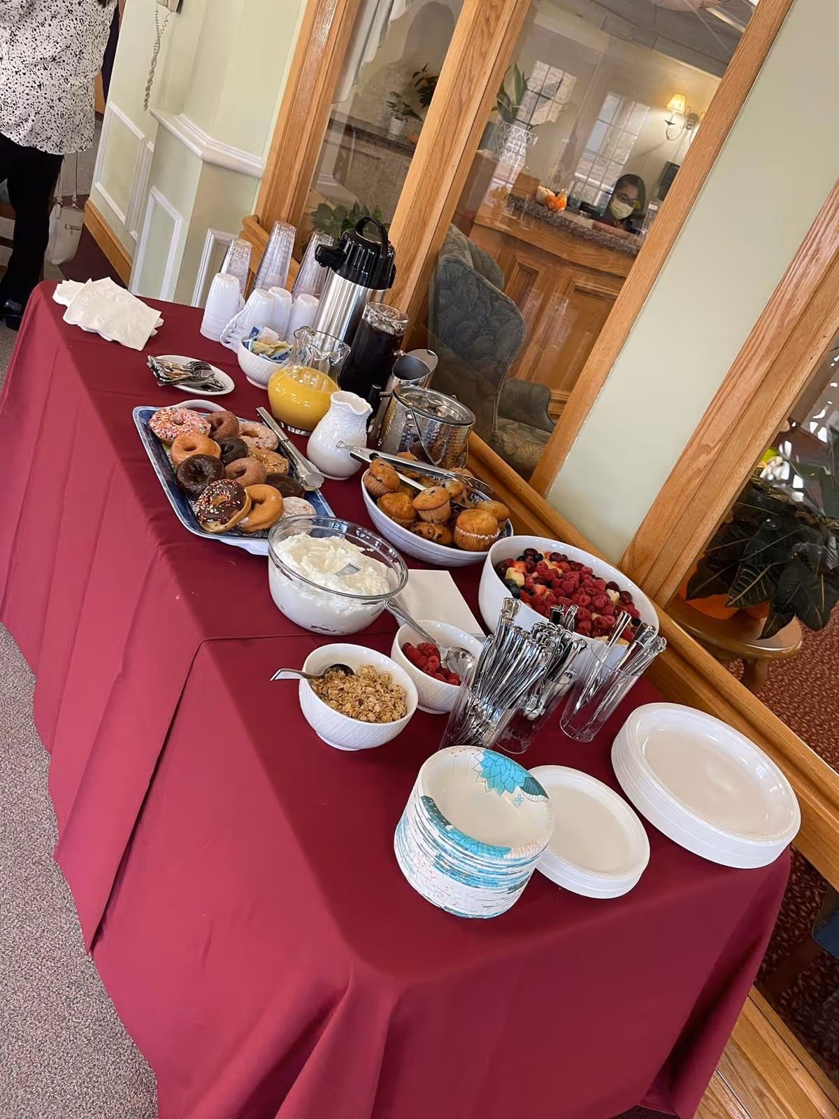 A buffet table covered with a red tablecloth displaying a variety of breakfast items including donuts, muffins, granola, yogurt, fresh berries, orange juice, coffee, and disposable plates and utensils. The table is set against a wall with large wooden-framed windows reflecting a seating area and a person wearing a mask.
