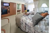 A well-lit bedroom in a senior living facility featuring a bed with patterned bedding and multiple pillows, a wooden coffee table with a flower vase, a beige armchair, a wall-mounted TV above a fireplace, and a window with sheer curtains allowing natural light to enter.
