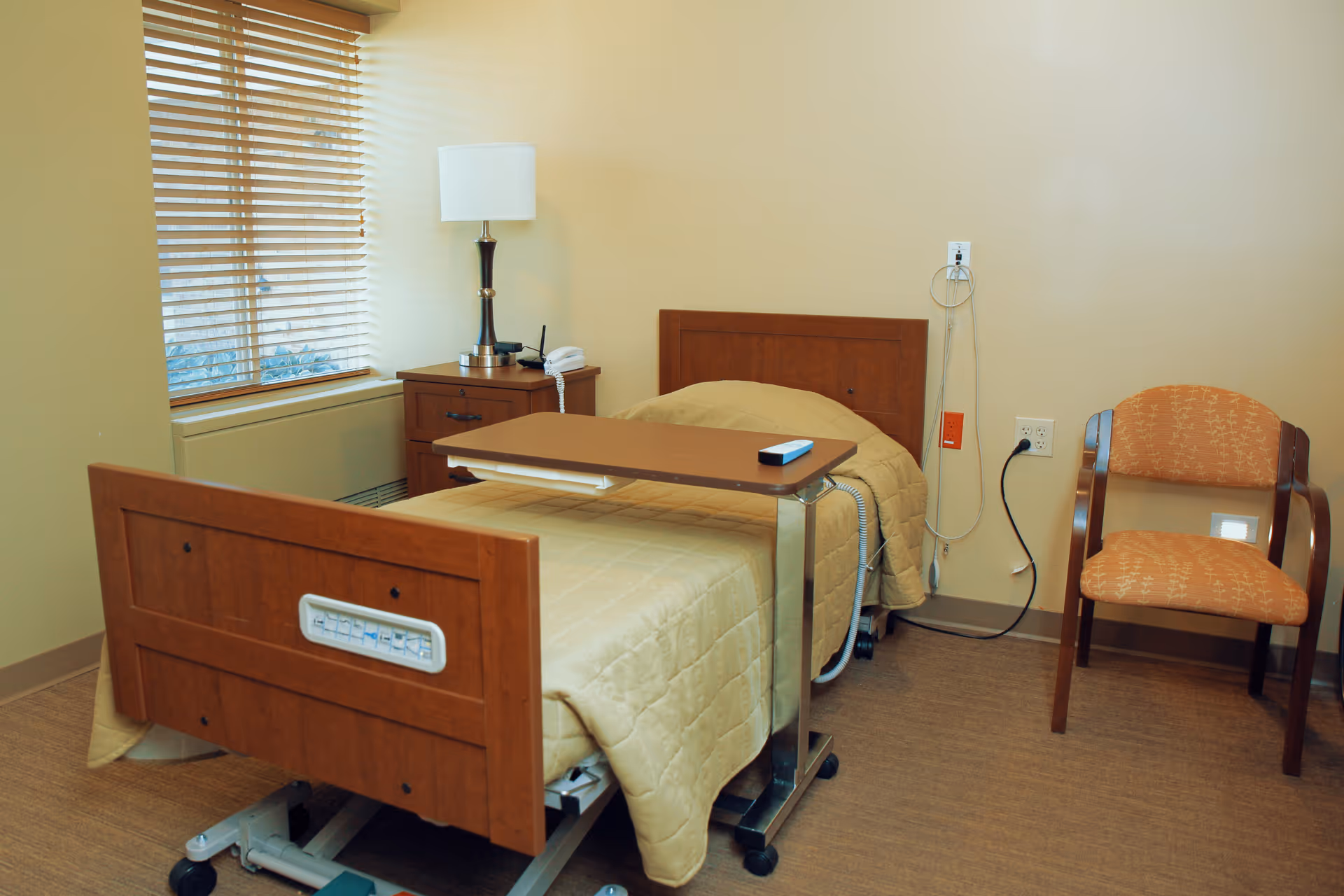 A small nursing facility bedroom with a single hospital bed covered in a beige quilt. There is a wooden nightstand with a lamp and a telephone next to the bed. A wooden chair with an orange patterned cushion is placed against the wall. A window with wooden blinds is on the left side of the room.